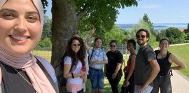 Das Selfie zeigt die Teilnehmer*innen bei einer Wanderpause, wo im Hintergrund der Chiemsee sichtbar ist.